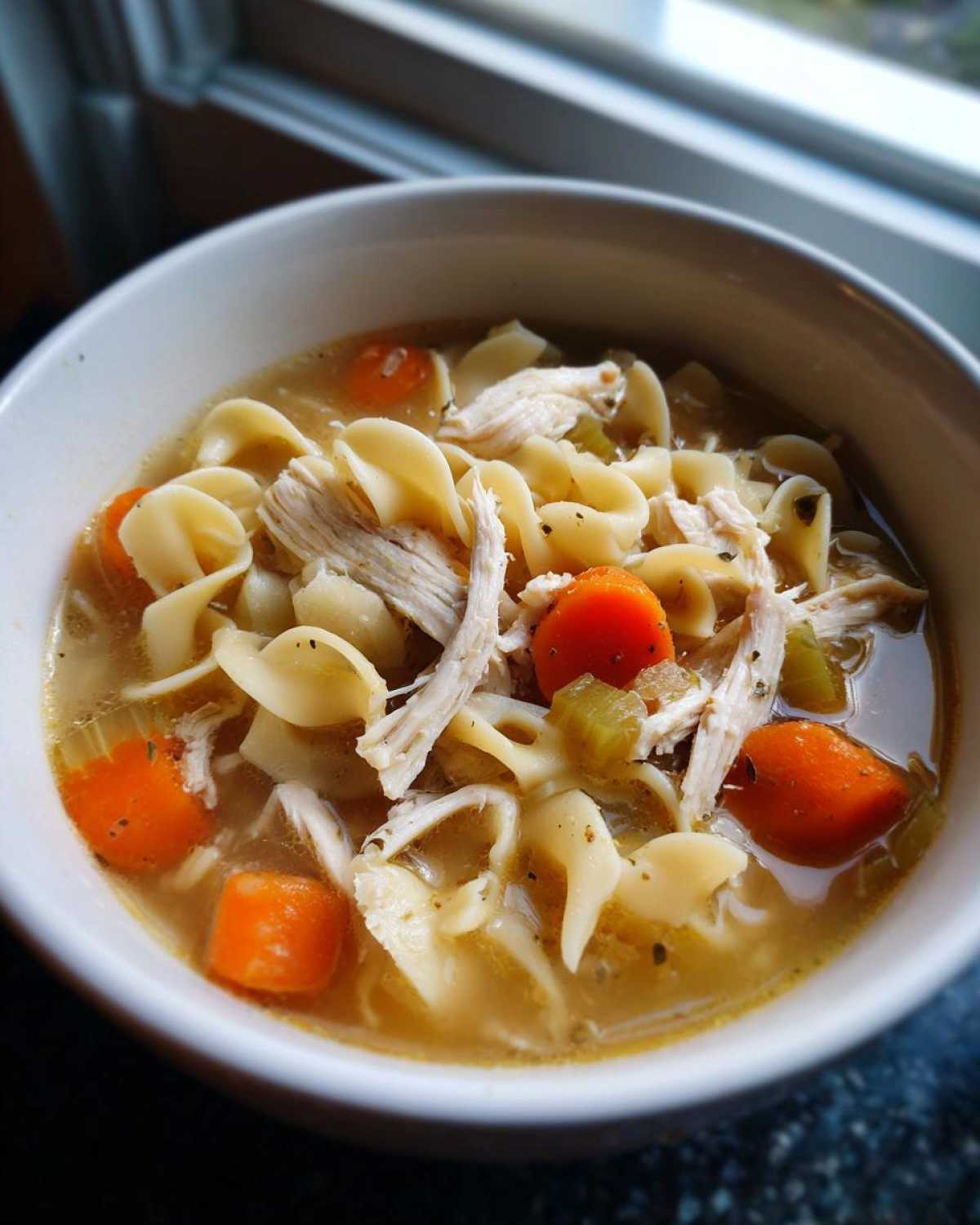 Close-up of a white bowl filled with homemade turkey noodle soup, featuring shredded turkey, carrots, and egg noodles.