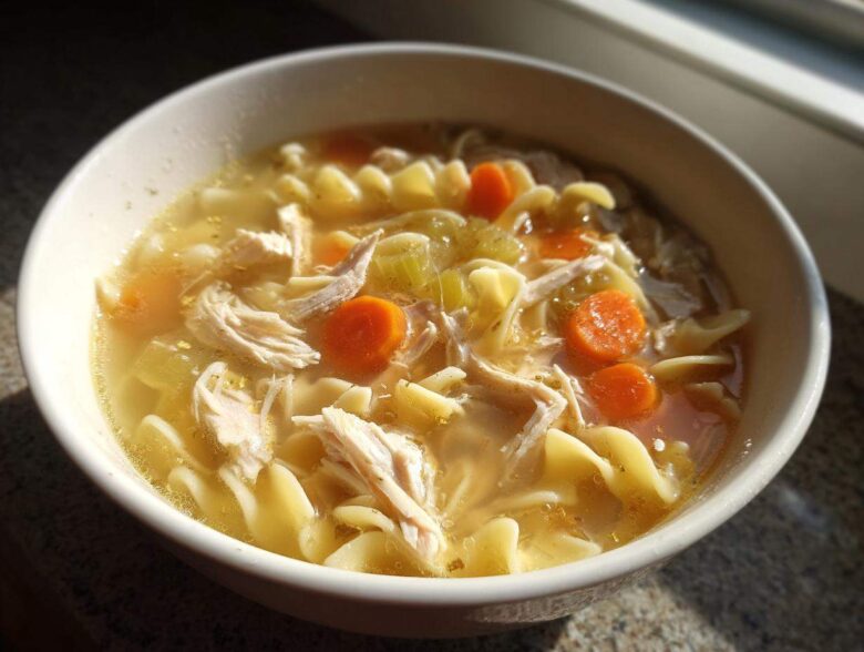 Close-up of a white bowl filled with steaming turkey noodle soup, featuring shredded turkey, egg noodles, and bright orange carrot slices.