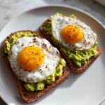 Two slices of perfect avocado toast topped with bright sunny-side-up eggs and chili flakes.