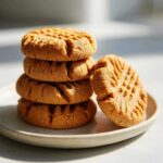 A stack of four golden brown 3-ingredient peanut butter cookies with classic fork marks, next to one leaning cookie on a small plate.