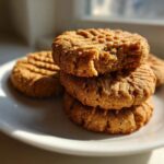 A stack of golden brown 3-ingredient peanut butter cookies on a white plate, one cookie on top has a bite taken out.