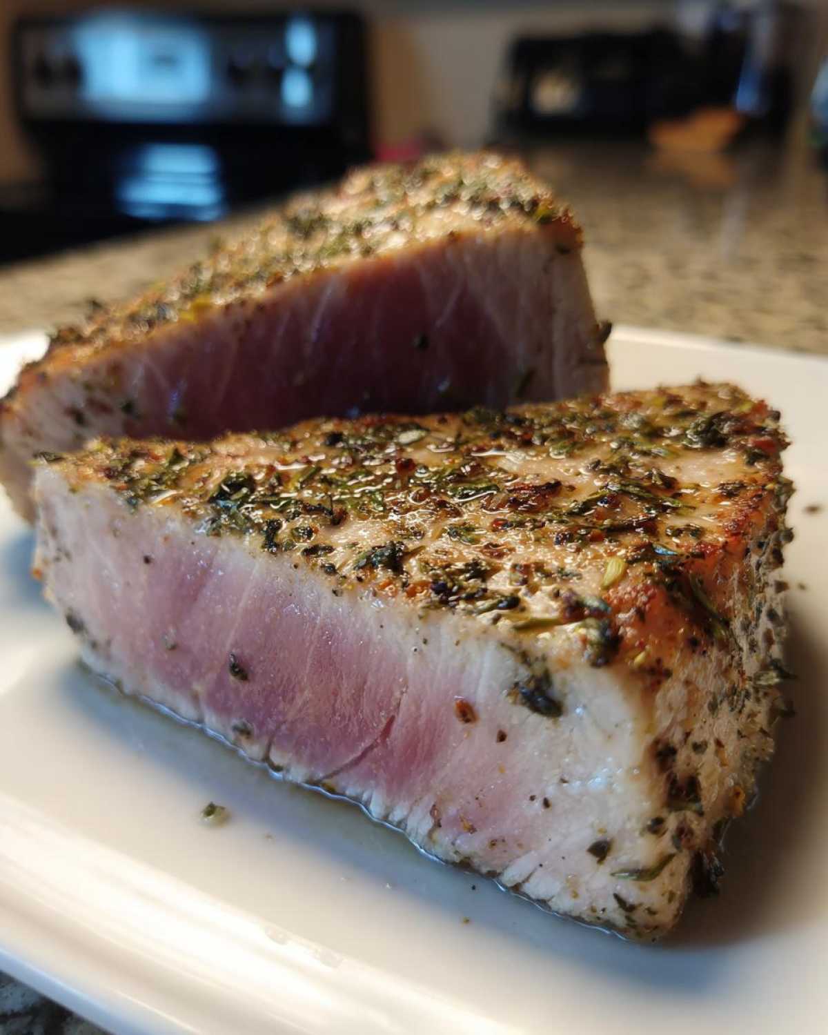 Two thick slices of medium-rare air fryer Tuna steak, heavily seasoned with herbs, resting on a white plate.