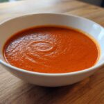 A close-up shot of rich, vibrant red Tomato Soup served in a white bowl on a wooden table.