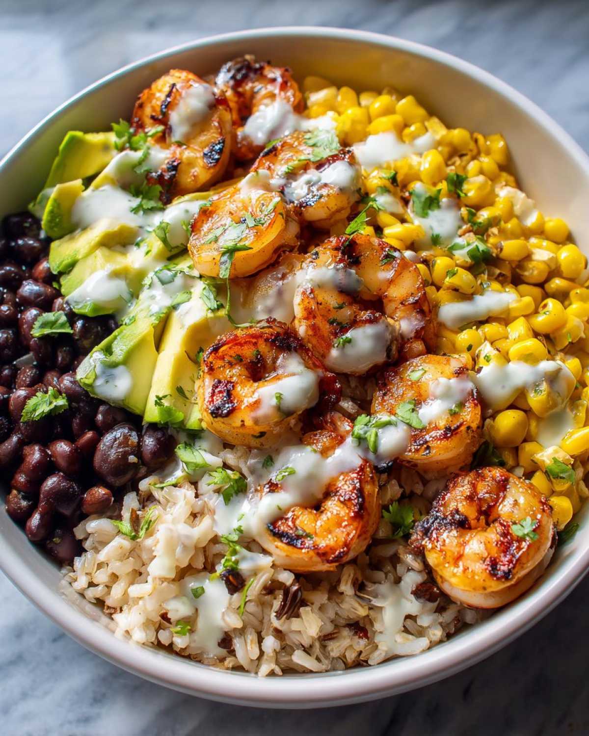 Close-up of a vibrant shrimp bowl featuring grilled shrimp, brown rice, black beans, corn, avocado, and a creamy white sauce.