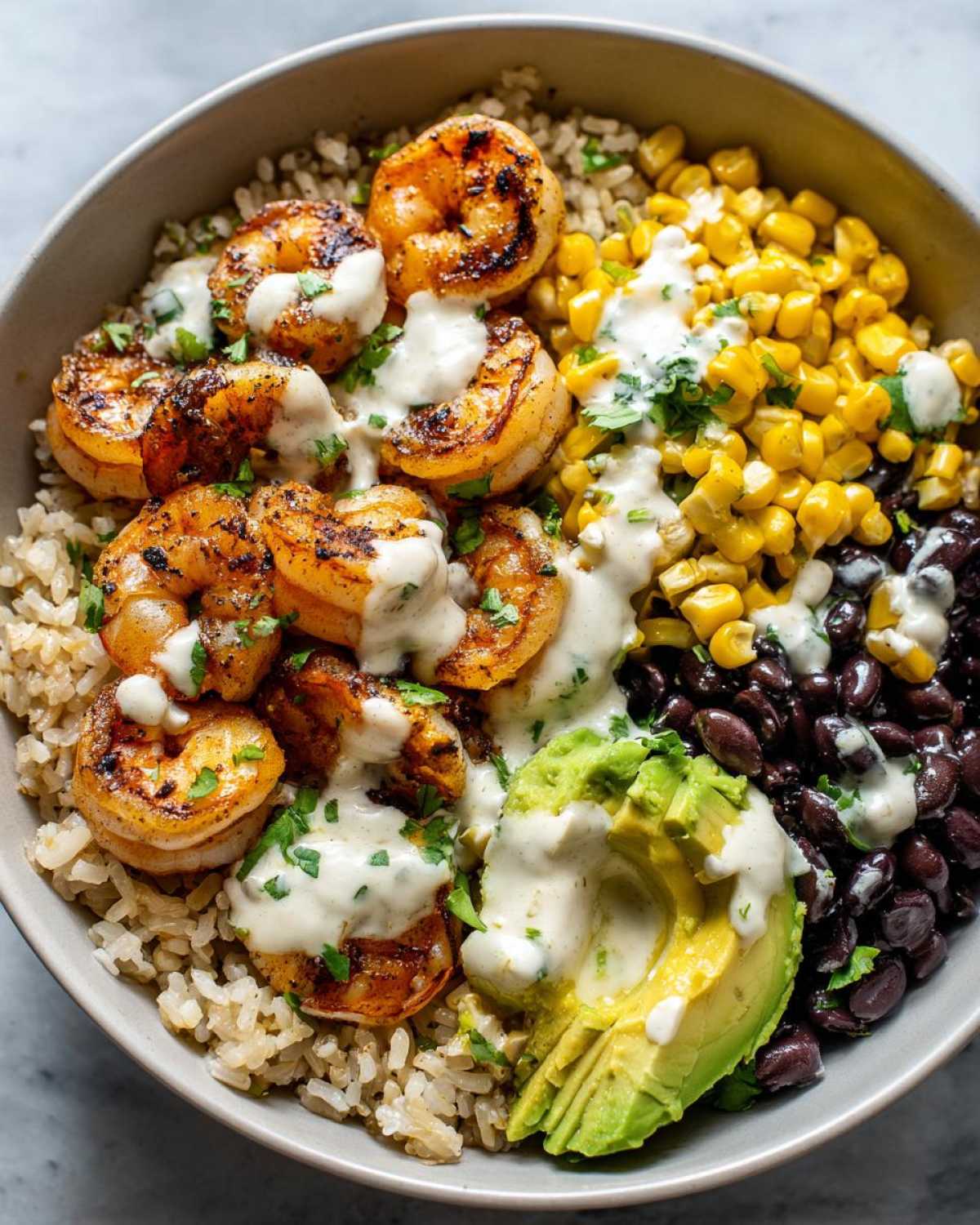 Close-up of a flavorful shrimp bowl featuring grilled shrimp, brown rice, black beans, corn, avocado, and a creamy white sauce.