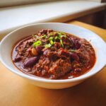 Close-up of a rich, dark red bowl of homemade chili filled with beef chunks and kidney beans, topped with green onions.