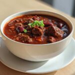A close-up of a white bowl filled with rich, dark red chili featuring chunks of beef and kidney beans, garnished with green onions.