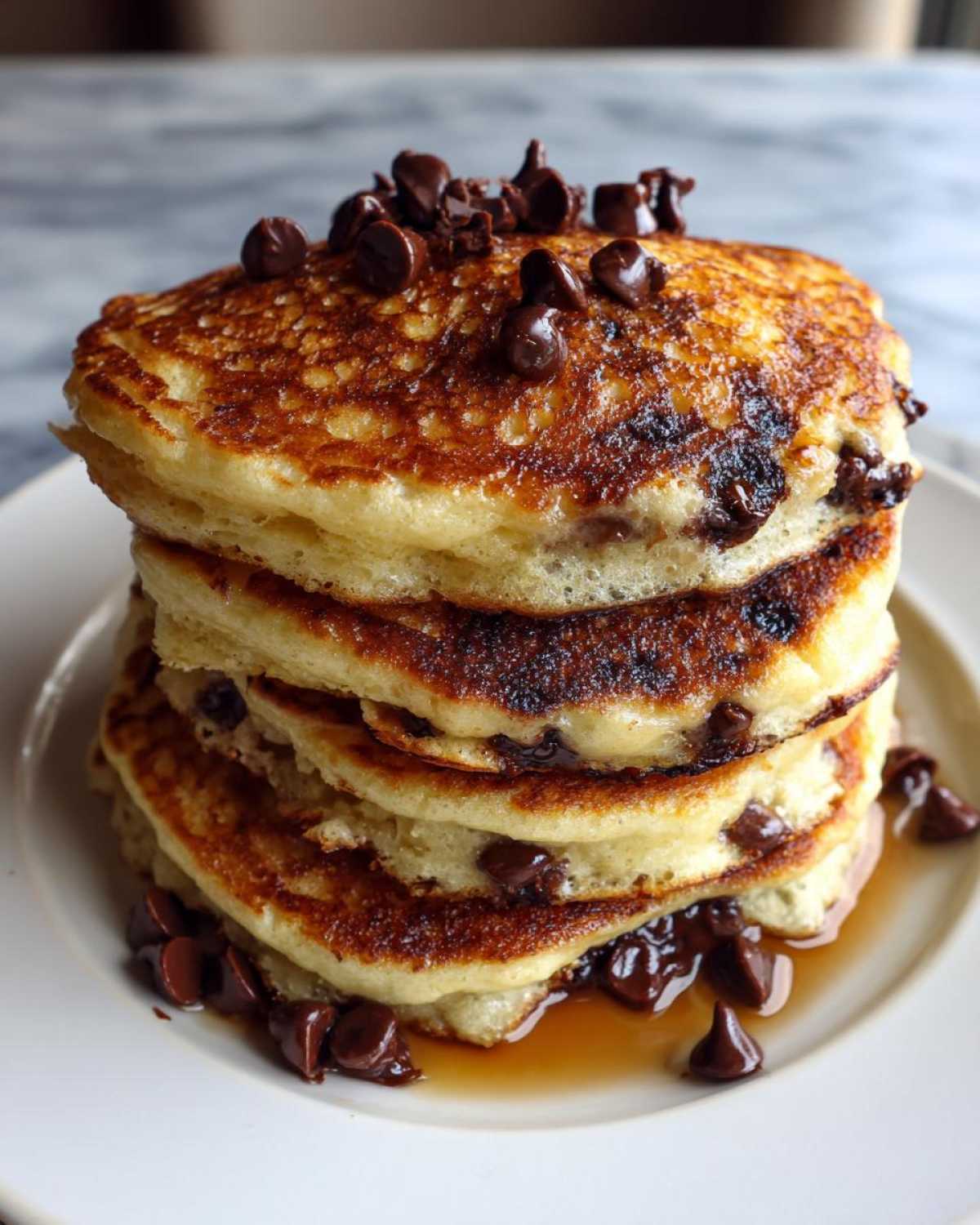 A tall stack of golden brown chocolate chip pancakes topped with melted chocolate chips and syrup.