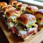 Close-up of several freshly baked grinder sliders loaded with melted cheese, salami, lettuce, and olives on a wooden board.
