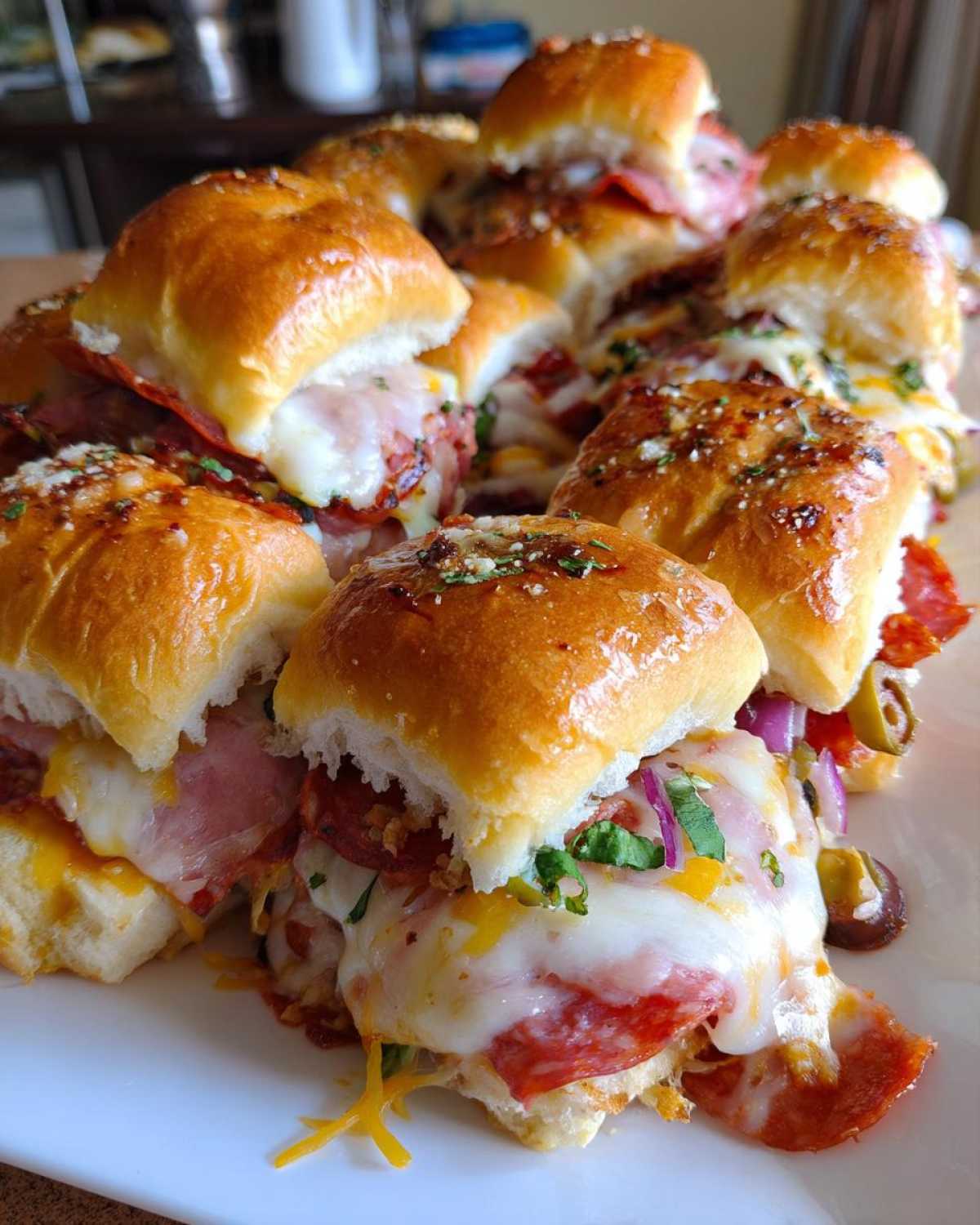 A pile of freshly baked, cheesy grinder sliders topped with herbs and glistening buns.