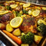 Close-up of roasted chicken thighs with crispy skin, potatoes, carrots, and broccoli, topped with lemon slices.