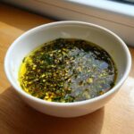 Close-up of a white bowl containing an amazing olive oil bread dip loaded with green herbs and chili flakes.