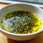 Close-up of a white bowl containing vibrant yellow olive oil bread dip topped with green herbs and red pepper flakes.