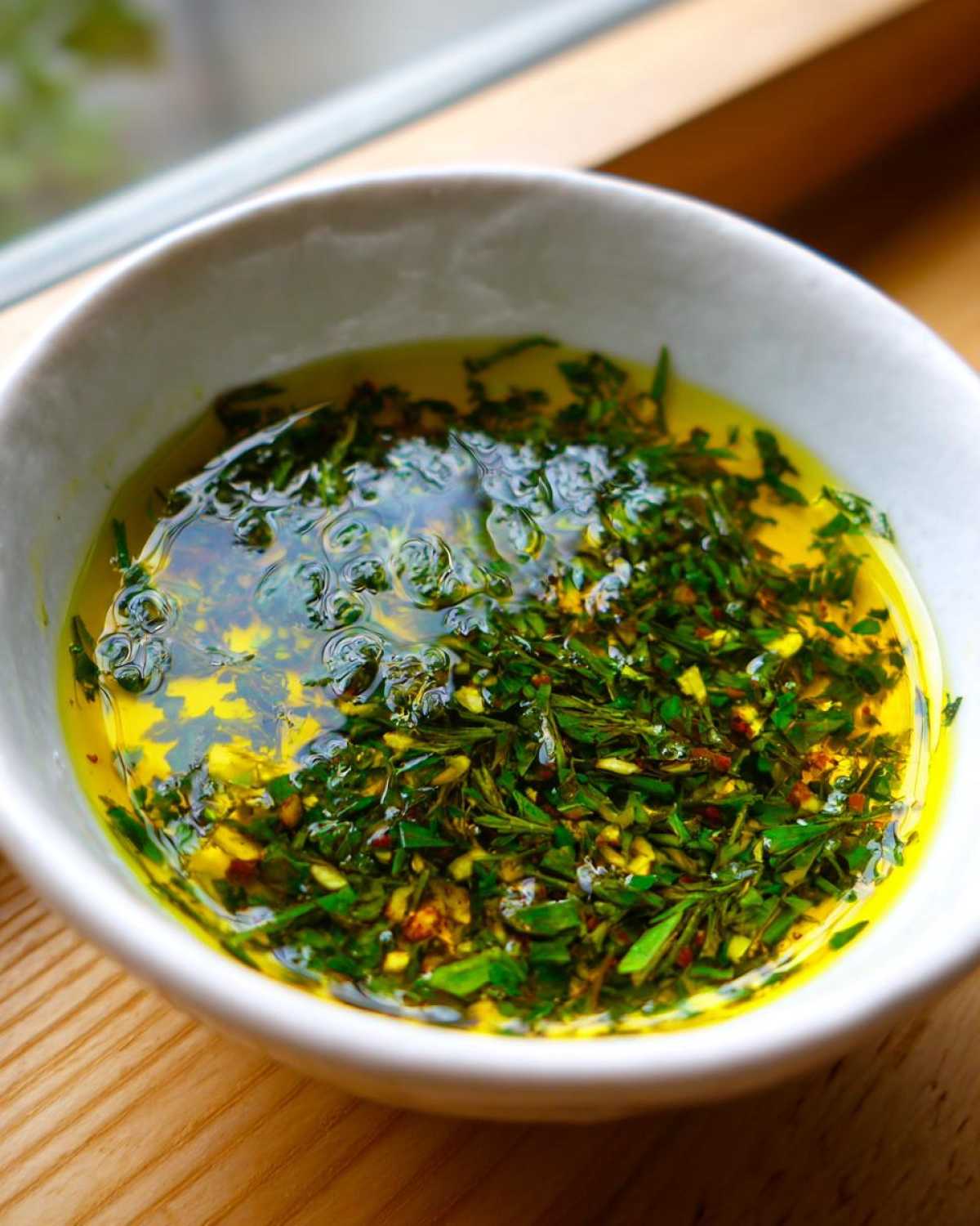 Close-up of a white bowl filled with bright yellow olive oil bread dip mixed with fresh green herbs and spices.