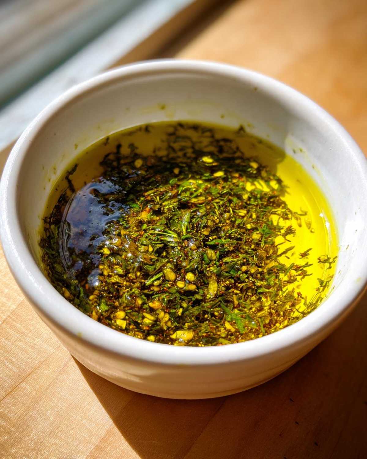 Close-up of a white bowl filled with bright yellow olive oil bread dip mixed with dark green herbs and spices.