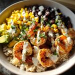 Close-up of a vibrant shrimp bowl featuring seasoned shrimp, brown rice, black beans, corn, avocado, and a creamy white sauce.