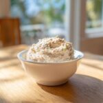 A creamy, textured bowl of homemade tuna dip garnished with small green pieces, sitting on a wooden table in bright sunlight.