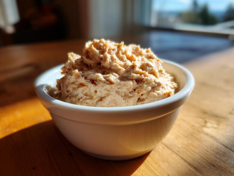 A close-up of a creamy, textured tuna dip served in a small white bowl on a sunlit wooden table.