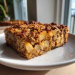 A close-up of a square slice of Apple Cinnamon Baked Oatmeal, showing visible chunks of cooked apple and a crunchy oat topping.