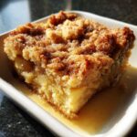 Close-up of a square slice of moist apple cobbler topped with cinnamon streusel, sitting in a pool of vanilla sauce.