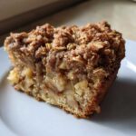 A close-up of a moist slice of apple crisp cake featuring chunks of apple and a thick, crunchy oat crumble topping.