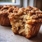 A close-up view of a bitten Apple oatmeal muffin showing the soft interior and cinnamon crumb topping, with another muffin blurred in the background.