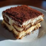 A close-up, beautifully lit slice of authentic Tiramisu showing layers of cream, coffee-soaked ladyfingers, and a heavy dusting of cocoa powder.