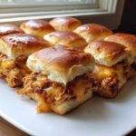 A close-up of baked bbq chicken sliders featuring shredded chicken, melted cheddar cheese, and glossy buns on a white platter.