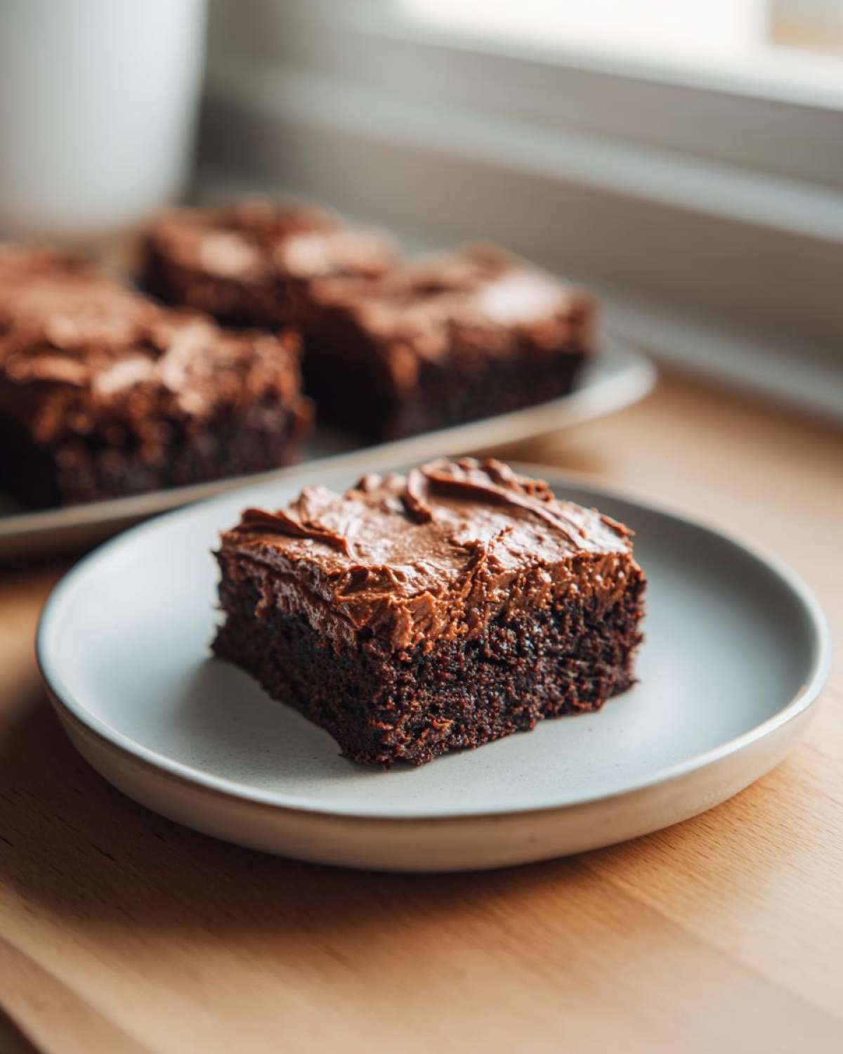 A close-up of a square slice of rich, fudgy banana bread brownies topped with thick chocolate frosting on a light plate.