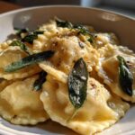 A close-up of glistening beef short rib ravioli served in a white bowl, topped with crispy sage leaves and brown butter sauce.