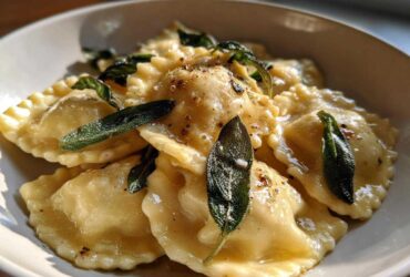 A close-up of glistening beef short rib ravioli served in a white bowl, topped with crispy sage leaves and brown butter sauce.