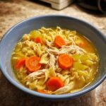 A close-up of a blue bowl filled with rich broth, egg noodles, shredded chicken, and bright orange carrot slices from the best chicken noodle soup.
