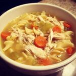 Close-up of a white bowl filled with steaming chicken noodle soup, featuring shredded chicken, egg noodles, carrots, and celery.