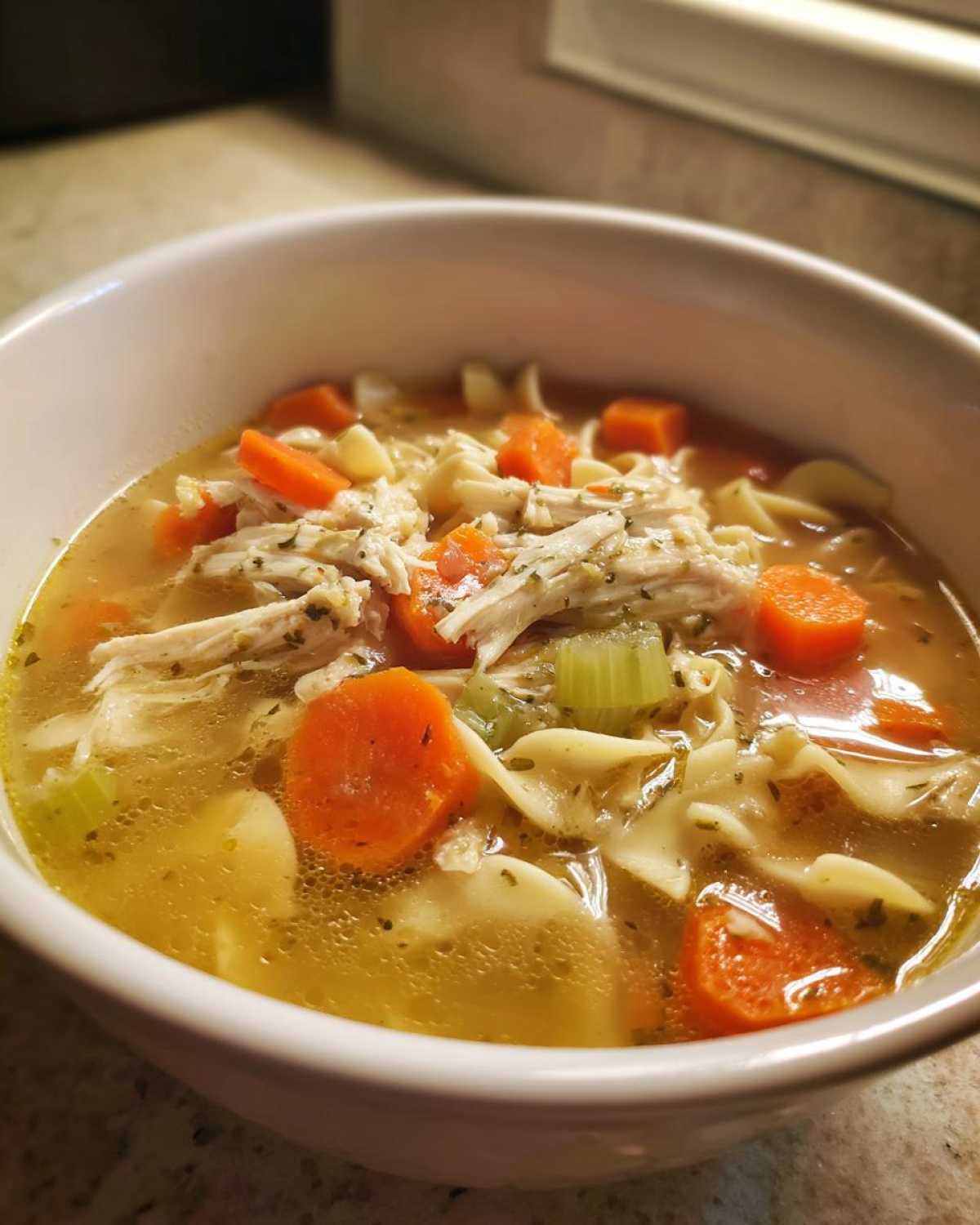 Close-up of a white bowl filled with homemade chicken noodle soup featuring shredded chicken, carrots, celery, and egg noodles.