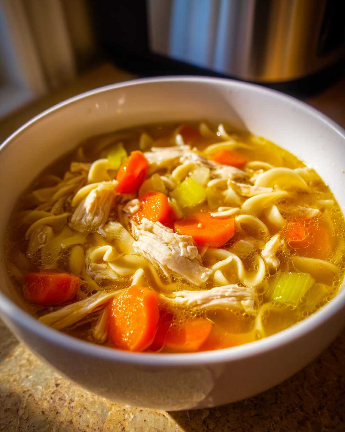 Close-up of a white bowl filled with rich broth, egg noodles, shredded chicken, carrots, and celery, ready to eat chicken noodle soup.