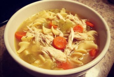 Close-up of a white bowl filled with steaming chicken noodle soup, featuring shredded chicken, egg noodles, carrots, and celery.