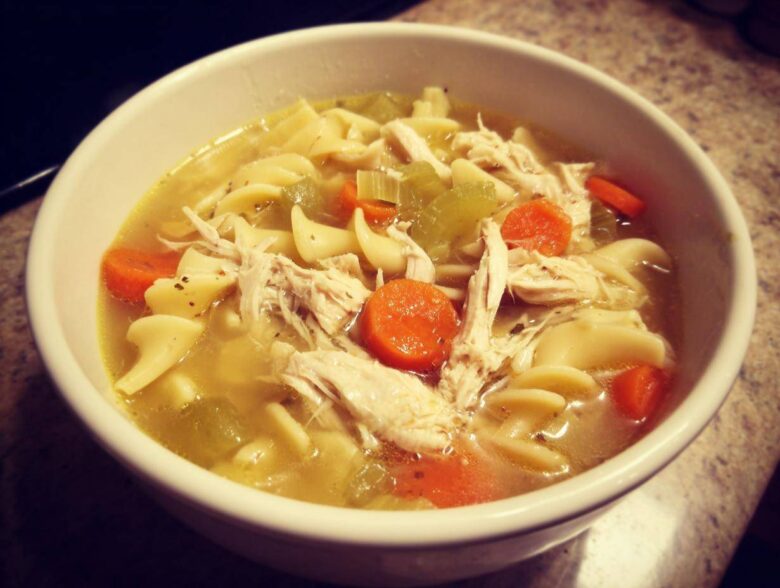 Close-up of a white bowl filled with steaming chicken noodle soup, featuring shredded chicken, egg noodles, carrots, and celery.