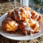 A close-up of several golden-brown baked apple fritters drizzled generously with white vanilla glaze.