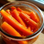 Close-up overhead shot of bright orange pickled carrots packed tightly in a clear glass jar, glistening in the light.