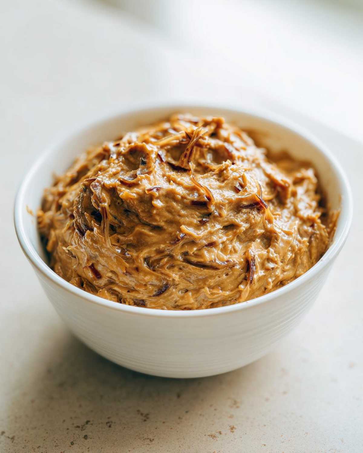 A close-up of thick, creamy caramelized onion dip swirled in a white bowl, showing visible strands of caramelized onions.