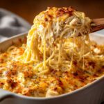 A wooden spoon lifts a cheesy, creamy portion of chicken spaghetti from a baked casserole dish, showing long cheese pulls.