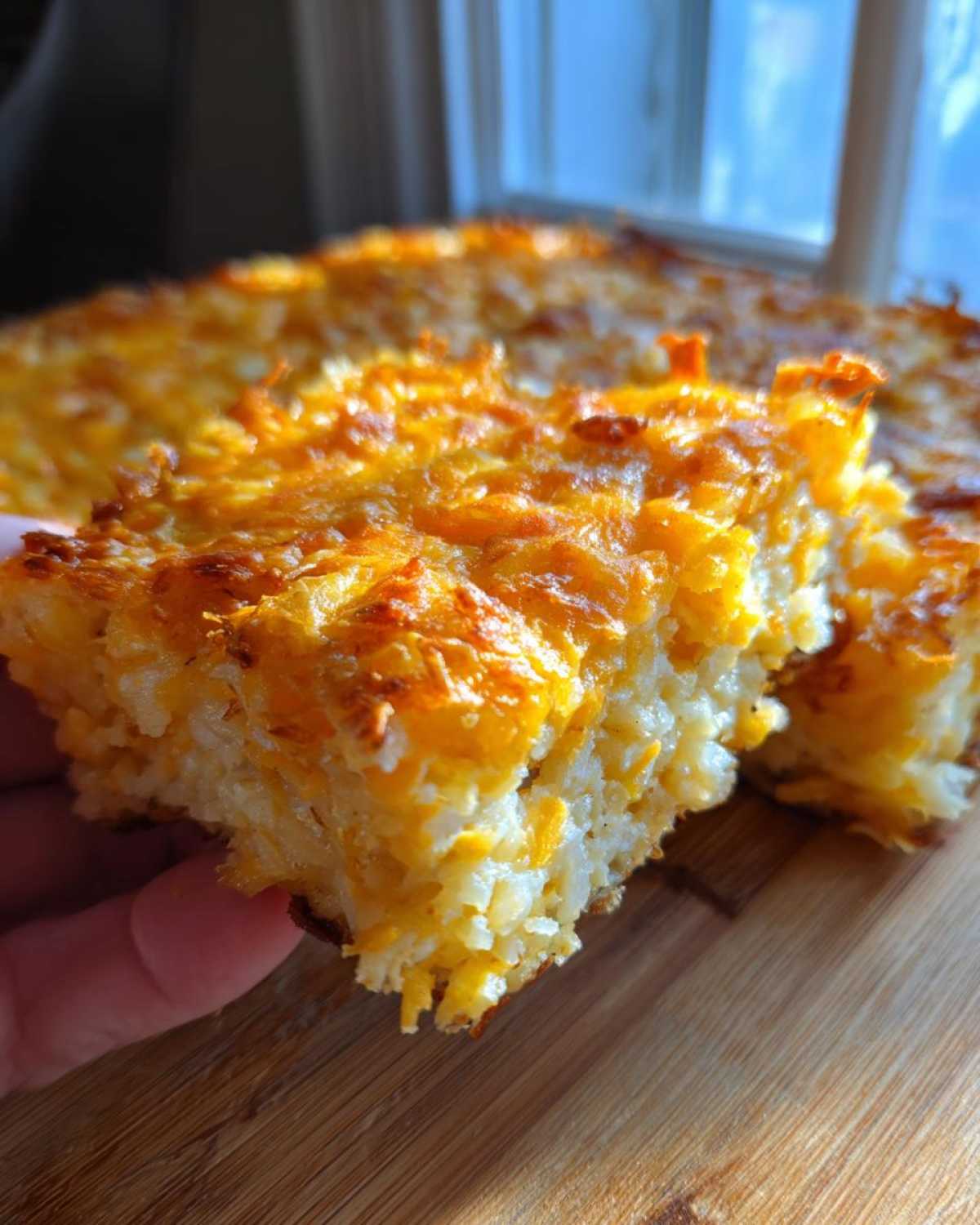 A hand holding a thick, square slice of hashbrown casserole, showing shredded potatoes and melted cheese.