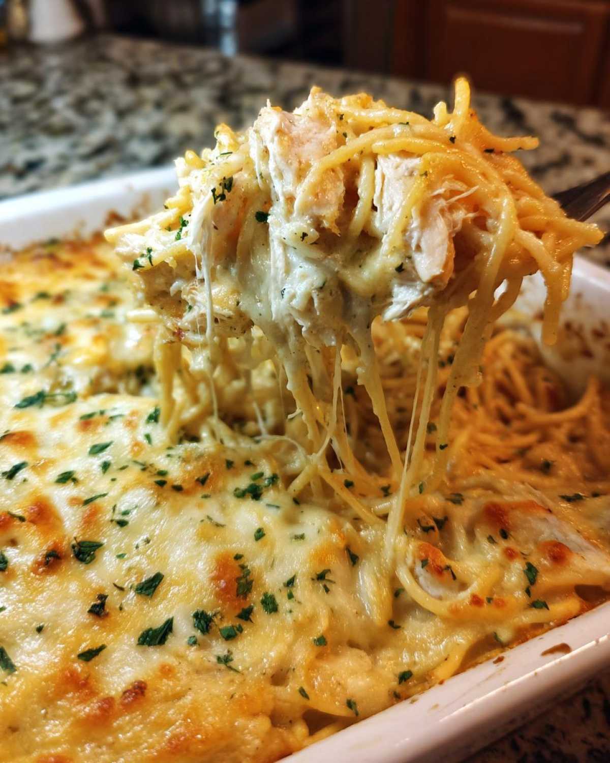 A fork lifts a cheesy, stringy portion of baked chicken spaghetti topped with melted cheese and parsley.