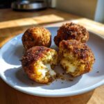 Four crispy, golden brown rice balls served on a white plate, one broken open showing the melted cheese filling inside.