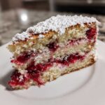 A close-up of a moist slice of cherry cake layered with bright red fruit filling and topped with powdered sugar.