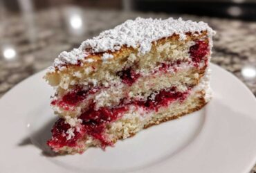 A close-up of a moist slice of cherry cake layered with bright red fruit filling and topped with powdered sugar.