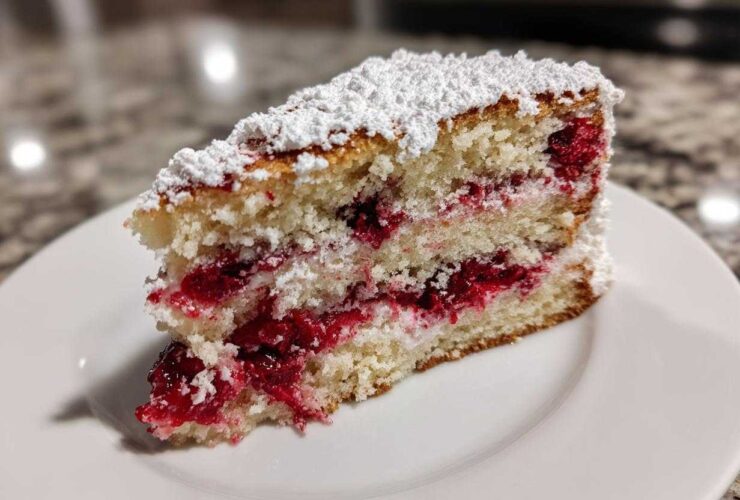 A close-up of a moist slice of cherry cake layered with bright red fruit filling and topped with powdered sugar.