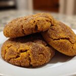 Three chewy pumpkin snickerdoodles coated in cinnamon sugar stacked on a white plate.