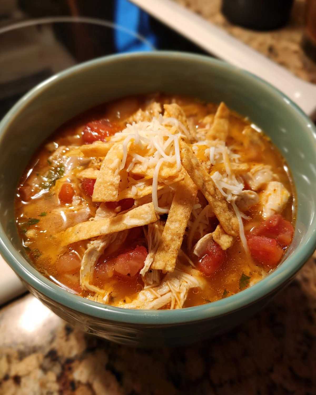 Close-up of a bowl of spicy chicken tortilla soup, filled with shredded chicken, tomatoes, and topped with crispy tortilla strips and shredded cheese.