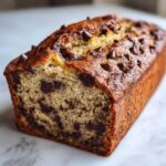 A close-up of a golden-brown chocolate chip banana bread loaf with visible chocolate chips throughout the moist interior.